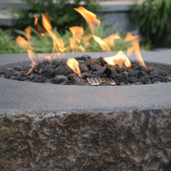 A close-up shot of a round fire pit with a textured gray surface and a prominent flame burning in the center. Dark lava rocks are visible beneath the flame.