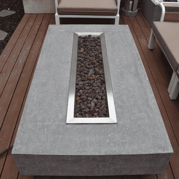  A bird's-eye view of a contemporary outdoor fire pit table. The table is rectangular and made of light gray concrete. A long, rectangular stainless steel burner is recessed into the center, filled with dark gray and brown lava rocks. Part of a wooden deck and blurred patio furniture are visible around the table.