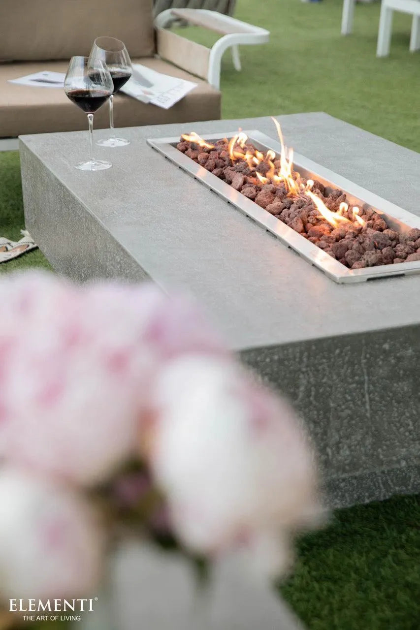 A rectangular concrete fire pit table with a stainless steel linear burner and flames sits on a green lawn with blurred pink flowers in the foreground. Two wine glasses filled with red wine are placed on the table.