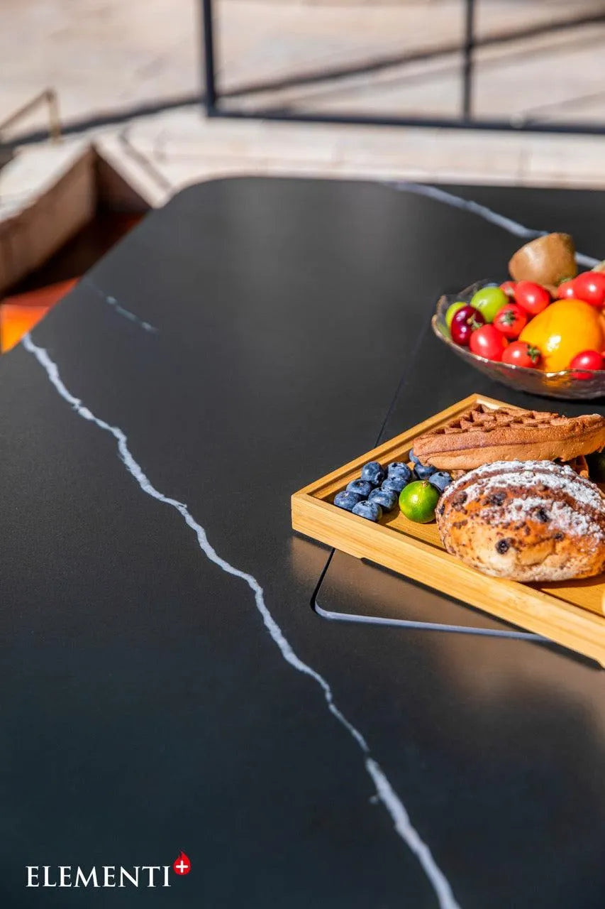 Close-up on a black table featuring a white veined pattern, with a wooden board holding bread and blueberries and a bowl of fruit. The "ELEMENTI+" logo is in the bottom left.