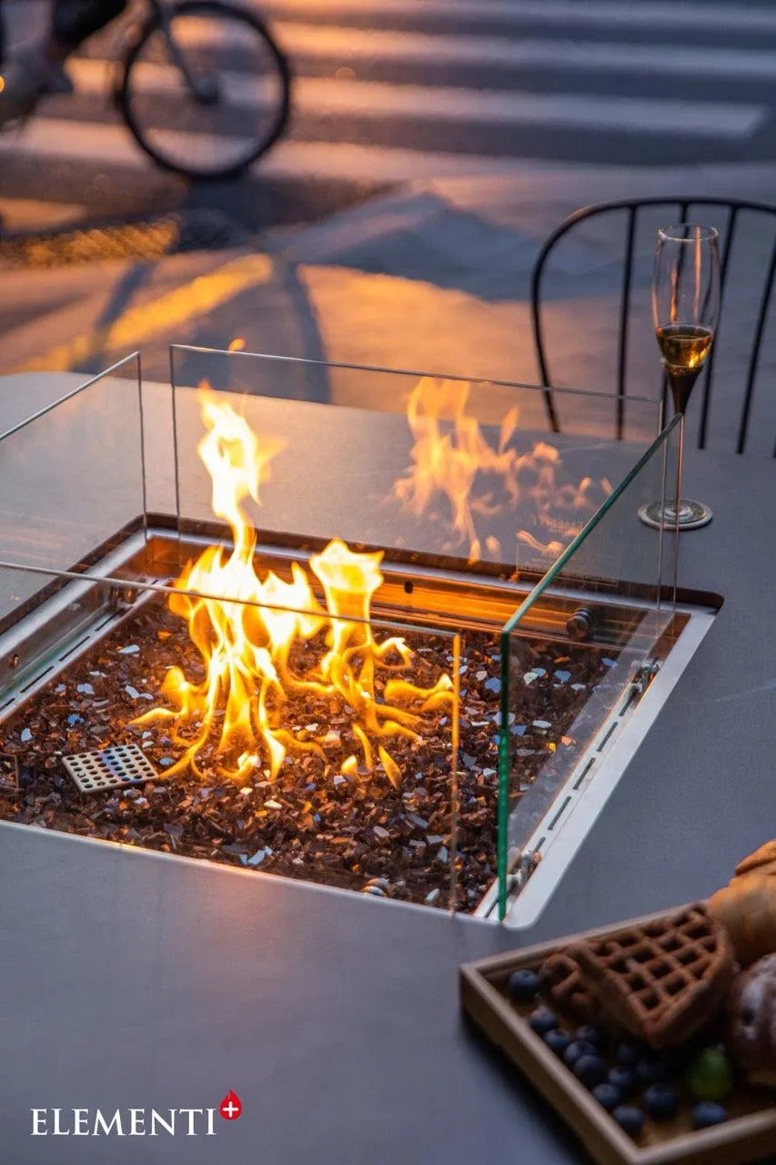 Close-up of a square outdoor fire pit table with a glass surround containing a lit fire. A champagne flute and a wooden tray with a waffle dessert are on the table. Dusk lighting with a blurred background. The "ELEMENTI+" logo is visible.