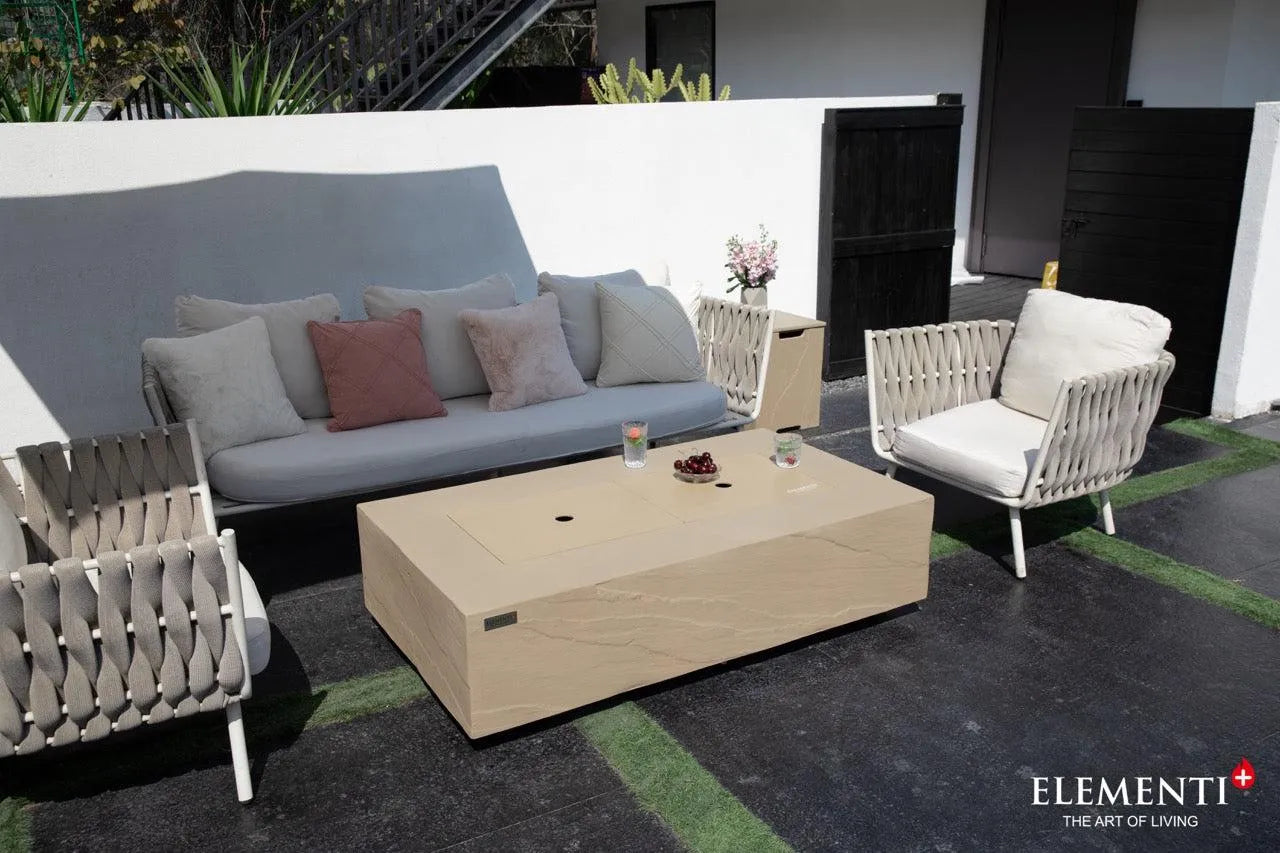 A rectangular concrete fire table with a metal cover sits on a patio with a gray sofa, decorative pillows, and two woven armchairs. A white wall and dark slatted doors are in the background.