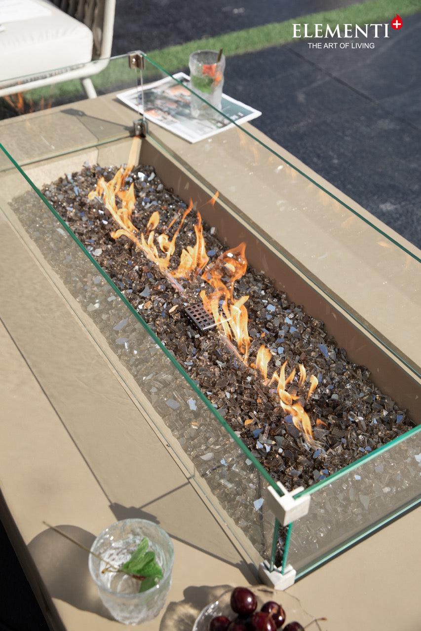 A rectangular concrete fire pit table with a glass wind guard and a linear flame burning on a bed of dark lava rock. Two glasses and a small dish are visible on the table.