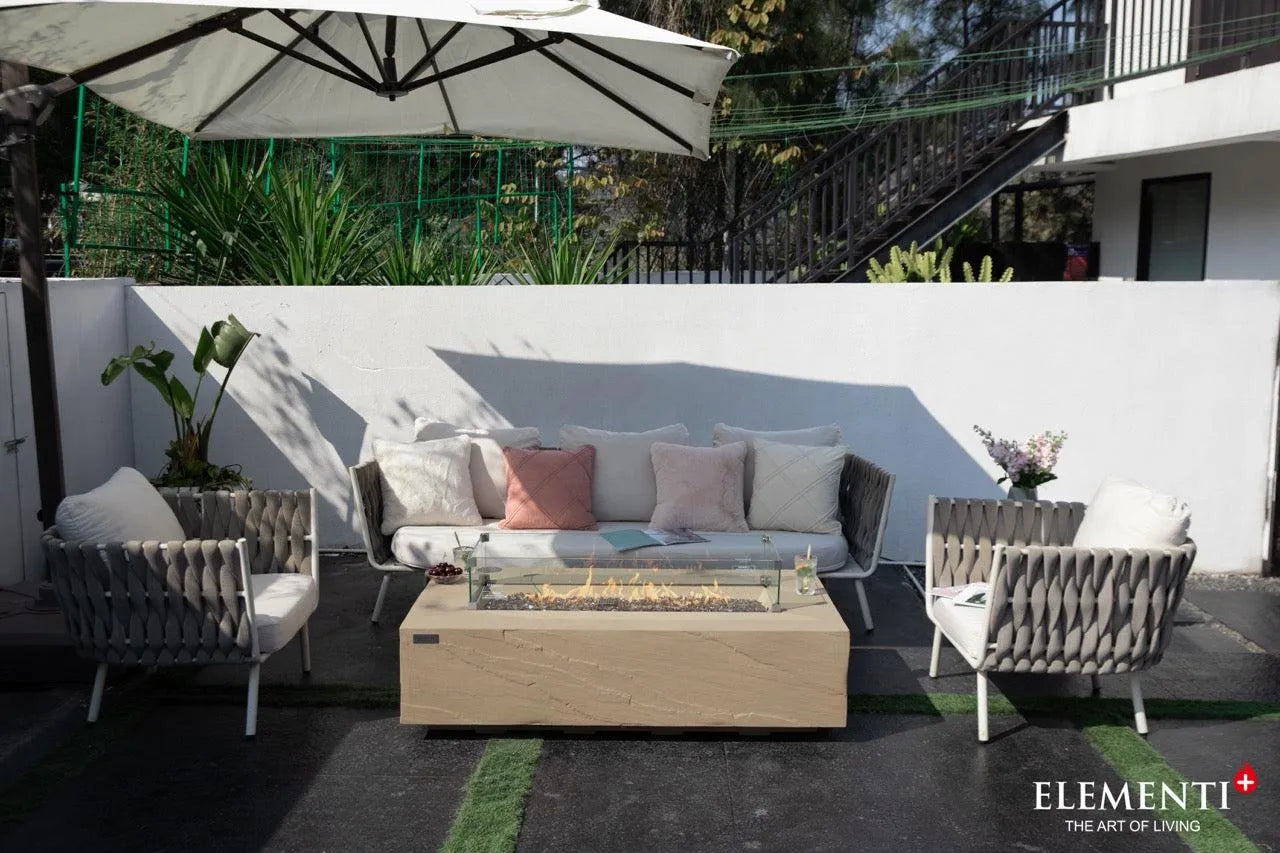 A rectangular concrete fire pit table with a glass wind guard and flames sits on a patio with a white sofa, decorative pillows, and two armchairs. A white umbrella provides shade, and a building with stairs is in the background.