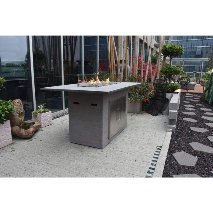 A modern, rectangular bar height fire pit table made of natural stone and concrete. The table features a hidden propane storage. A fire is lit protected by a glass wind screen that is around it.