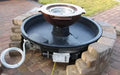 A black, concrete fire pit with a stainless steel gas burner in the center. Flames are visible burning on the water's surface. The fire pit is surrounded by a shallow, black basin with a scupper design featuring four water spouts flowing down the sides