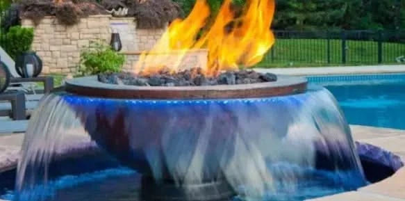 A round, stainless steel fire pit with a water basin. The fire pit has a black fire bowl with a flame burning in the center. The water basin is made of black plastic and has a square waterfall feature on one side.