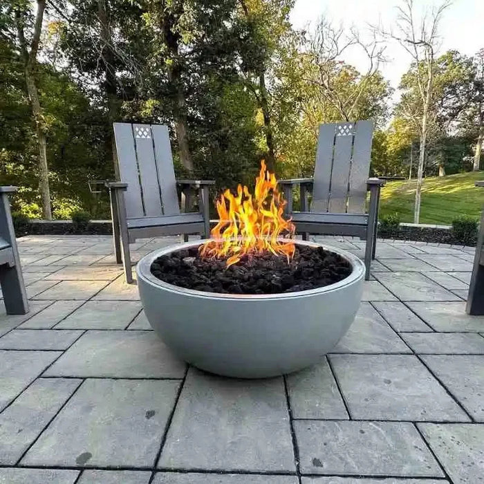 A round, stainless steel gas fire pit with a black powder-coated finish sits on a stone patio. Two black metal chairs with gray cushions flank either side of the fire pit. In the background, there is a green hedge and a brown wooden fence.