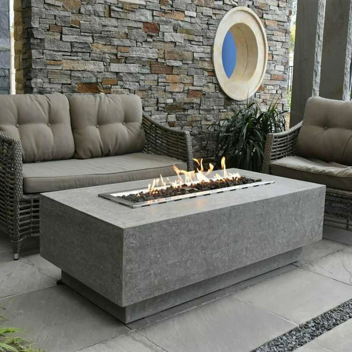 A rectangular fire pit made of dark concrete sits on a light colored concrete table. The fire pit has a rectangular stainless steel burner in the center with flames coming out of it.