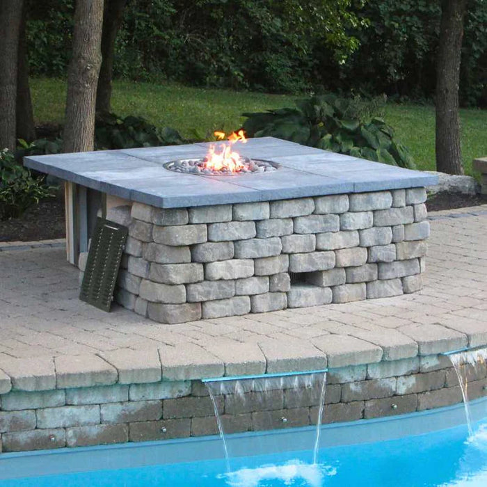 A square fire pit made out of light colored stone sitting on a brick wall next to a blue swimming pool. The fire pit has a stainless steel burner and flames coming out of it. There are lounge chairs and a small table on the patio behind the fire pit.