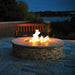 A stainless steel fire pit with a lit flame burning in the center. The fire pit is on a slate patio with a wooden bench nearby. There are trees and landscaping in the background, with a lake in the distance.