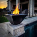 A square, hammered copper fire pit with a stainless steel burner pan in the center. The fire pit is sitting on a concrete base next to a swimming pool.