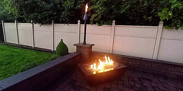 Hampered copper fire pit with a rectangular stainless steel burner and flames burning in the center. The fire pit has a decorative grate on top. Rectangular hammered copper fire pit with flames and a decorative grate. The fire pit rests on a black base.  