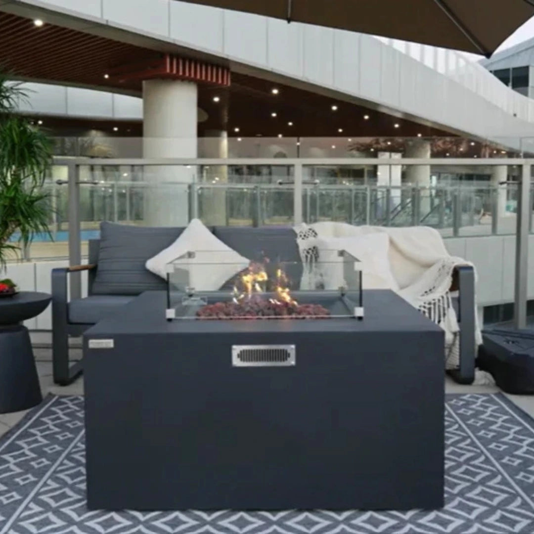 Lit fire pit with a glass wind guard on a patio with a glass railing, a gray sofa with pillows, and a patterned rug.