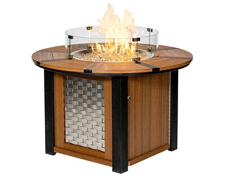 A round, gas-powered fire pit table with a glass flame guard. The table has a woven metal panel on the bottom half and a light brown top. The flames are visible through the glass guard.