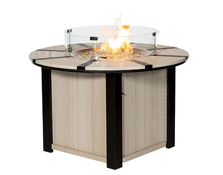 A round, wooden fire pit table with a glass flame guard and a flame burning inside. The table has a square base with black accents.