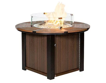  A round, wooden fire pit table with a black metal frame. The table has a glass wind guard and a flame burning inside.