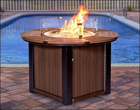 A round fire pit table with a glass flame guard and a flame burning inside. The table is made of brown poly lumber with a black metal frame. It is placed on a brick patio next to a swimming pool.