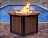 A round fire pit table with a glass flame guard and a flame burning inside. The table is made of brown poly lumber with a black metal frame. It is placed on a brick patio next to a swimming pool.