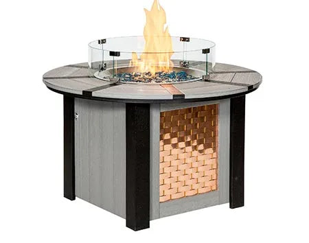 A round, gas-powered fire pit table with a glass flame guard. The table has a woven copper panel on the bottom half and a light gray top. The flames are visible through the glass guard.