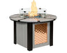 A round, gas-powered fire pit table with a glass flame guard. The table has a woven copper panel on the bottom half and a light gray top. The flames are visible through the glass guard.