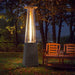 A silver pyramid-shaped propane patio heater with a woven base, casting a warm glow over a wooden bench and glasses on a table in a backyard at night.