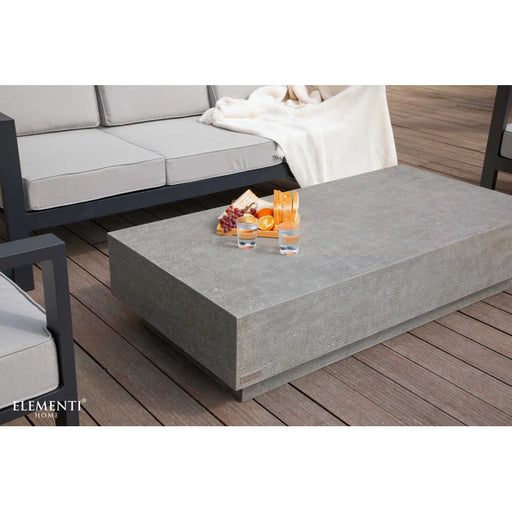 A large rectangular concrete coffee table in a space grey finish from the Elementi Home Tevere series, situated on a wooden patio next to outdoor seating with grey cushions.