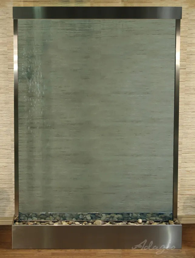 
A large, rectangular Adagio Mega Grandeur free standing Water Feature, featuring a clear glass water panel framed by brushed stainless steel. Water cascades down the glass, creating a textured, rippling effect, especially visible on the left side. 