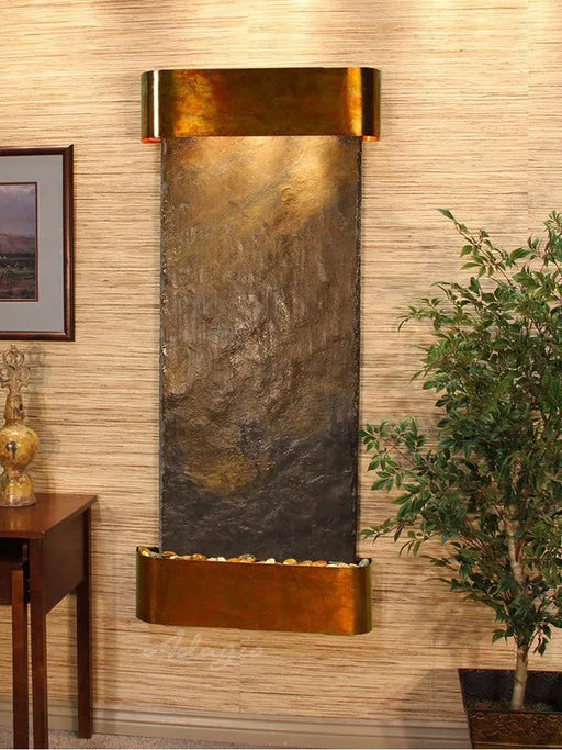 Wall-mounted water feature with bronze finish in a room with a plant and side table.