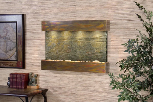 
A stunning Adagio Reflection Creek wall-mounted water fountain with an antique copper frame and a textured green slate face. Warm lights illuminate the water flowing over the slate, which has subtle horizontal striations, into a basin filled with decorative river rocks. The fountain is set against a textured, light beige wall, with a framed map visible to the left and green foliage on the right, suggesting an elegant indoor setting.
