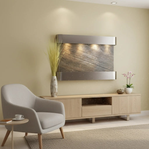 An Adagio Inspiration Falls wall-mounted water feature in a modern living room. The fountain has a silver frame and a textured, gray-brown stone surface with water flowing down it. It is mounted on a light tan wall above a light wood media console. To the left is a cream-colored armchair and a small side table with a cup on it. A tall vase with dried grasses sits on the left side of the media console, and a potted orchid is on the right. A large window with sheer white curtains is on the right side of the r
