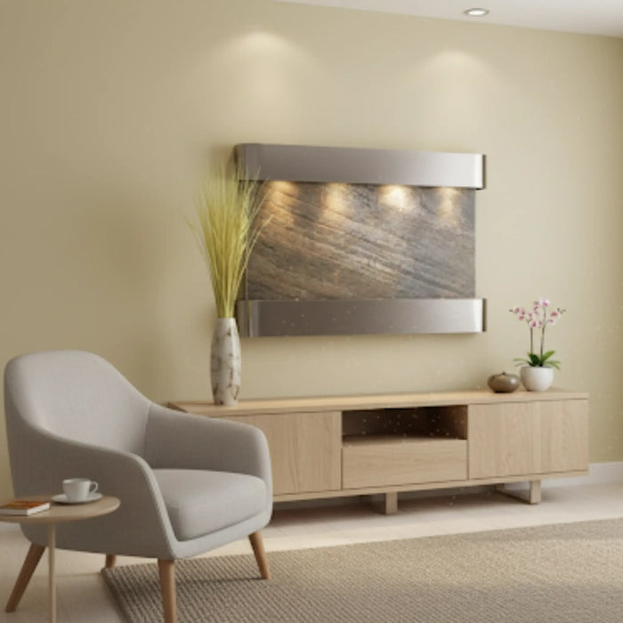 An Adagio Inspiration Falls wall-mounted water feature in a modern living room. The fountain has a silver frame and a textured, gray-brown stone surface with water flowing down it. It is mounted on a light tan wall above a light wood media console. To the left is a cream-colored armchair and a small side table with a cup on it. A tall vase with dried grasses sits on the left side of the media console, and a potted orchid is on the right. A large window with sheer white curtains is on the right side of the r