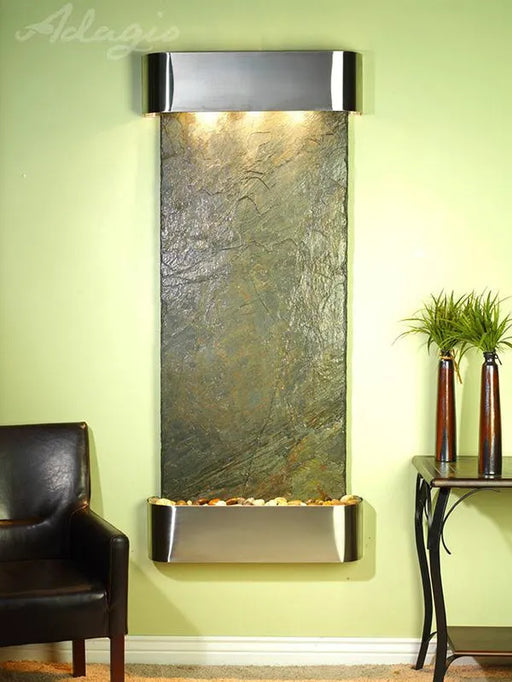 Wall-mounted water feature with decorative stones against a light green wall, next to a black armchair and small table with plants.
