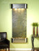 Wall-mounted water feature with decorative stones against a light green wall, next to a black armchair and small table with plants.
