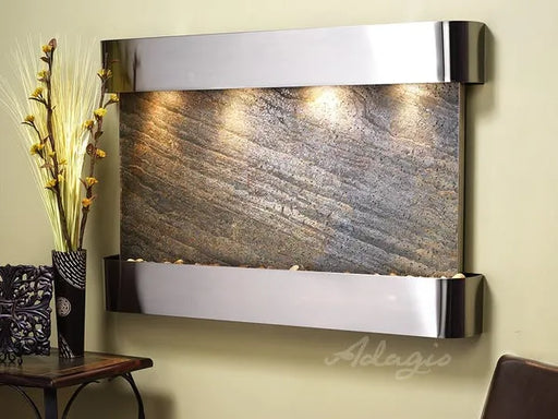 A close-up of an Adagio Inspiration Falls wall-mounted water feature with a silver frame and a textured, gray-brown stone surface. Water flows down the stone, which is illuminated by three lights from the top. Small decorative stones are visible in the basin at the bottom. To the left, a tall vase with dried yellow decorative branches sits on a dark wooden table next to a patterned dark vase. The wall behind the fountain is a light tan color.
