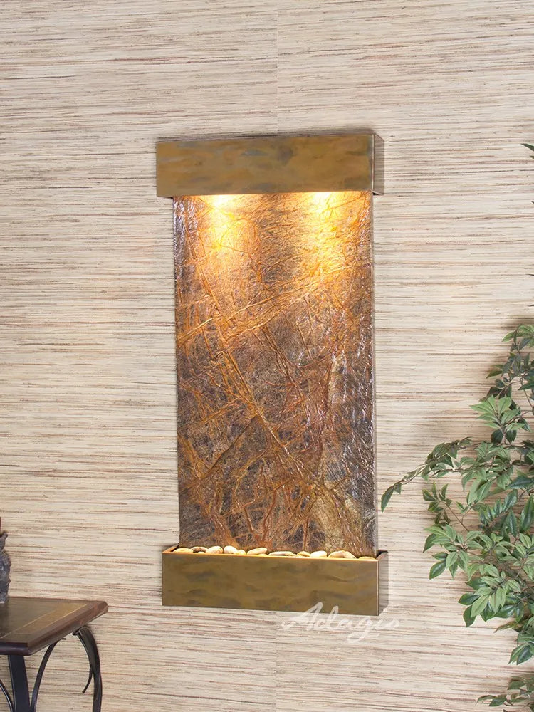 
A beautiful Adagio Whispering Creek wall-mounted water fountain with a warm copper frame and textured natural slate face. Warm lights illuminate the water cascading over the slate, which has prominent natural veining, into a basin filled with river rocks at the bottom.