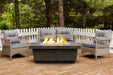 A cozy outdoor seating arrangement with a rectangular fire pit table as the centerpiece. The table is surrounded by two wicker chairs with gray cushions and overlooks a wooden deck with a white picket fence in the background.