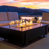 A rectangular dark gray fire table with glass wind guards and flames burning in the center, positioned on a patio with two light-colored sofas. A sunset sky and silhouetted mountains are in the background.
