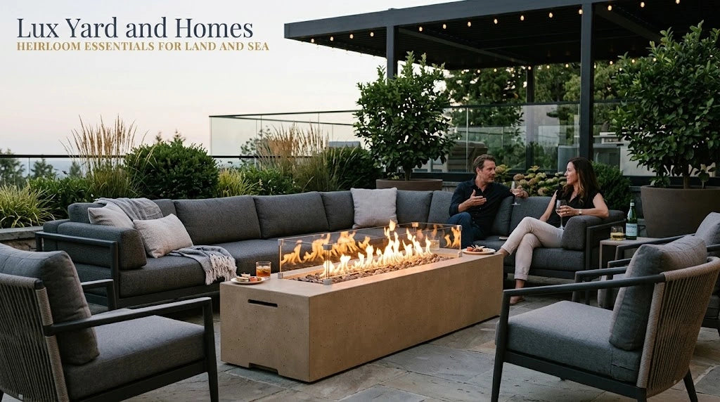Low profile American Fyre Designs Milan 72 inch outdoor natural gas fire table in textured Sedona sand concrete, operating with a linear flame and glass wind guard on a stone patio with deep seating sectional sofa, guests, and Lux Yard and Homes branding.