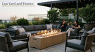 Low profile American Fyre Designs Milan 72 inch outdoor natural gas fire table in textured Sedona sand concrete, operating with a linear flame and glass wind guard on a stone patio with deep seating sectional sofa, guests, and Lux Yard and Homes branding.