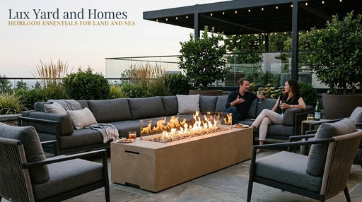 Low profile American Fyre Designs Milan 72 inch outdoor natural gas fire table in textured Sedona sand concrete, operating with a linear flame and glass wind guard on a stone patio with deep seating sectional sofa, guests, and Lux Yard and Homes branding.