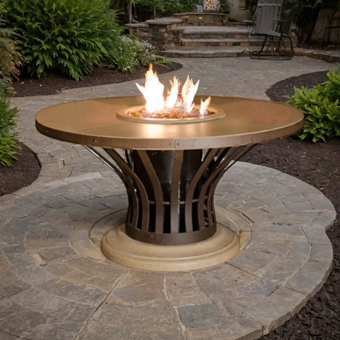 A photo of the American Fyre Designs Fiesta Round Dining Fire Table in a backyard setting. The fire table is lit, with a warm glow illuminating the surrounding patio furniture and creating a cozy atmosphere.