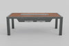 An angled front view of a bar-height outdoor grill table featuring a wood-slat top, a long built-in grill, and dark grey metal legs. A control panel with knobs is visible on the left side of the table, and two rectangular compartments are underneath.