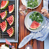 Overhead view of grilled watermelon slices on a grill and a plate of salad and watermelon on a wooden table
