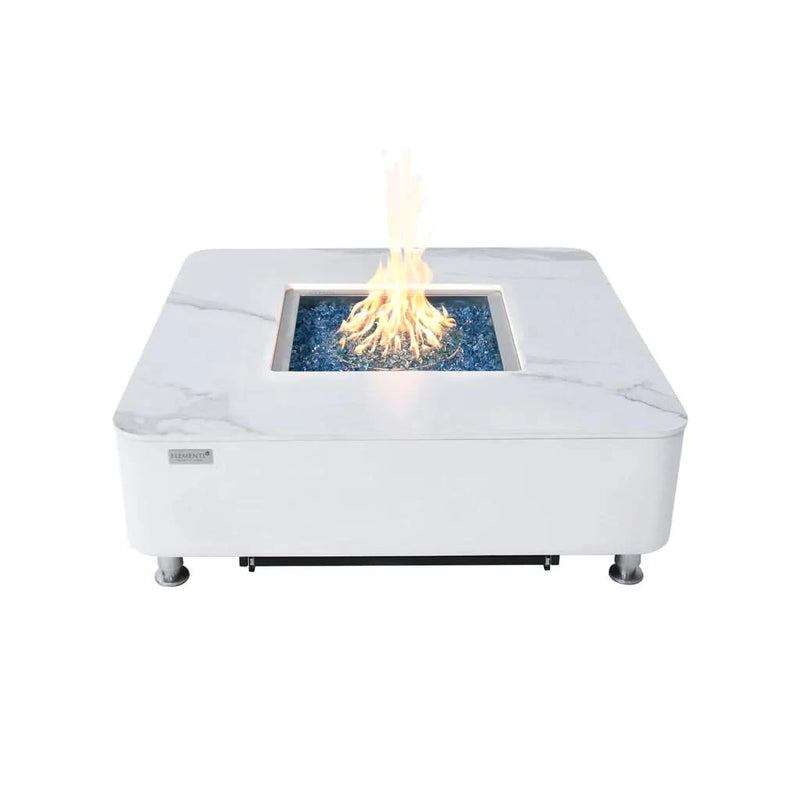 Modern white square fire pit table with a marble-look top and a stainless steel burner, lit with a flickering flame. The table has a white base and is elevated on small silver feet.