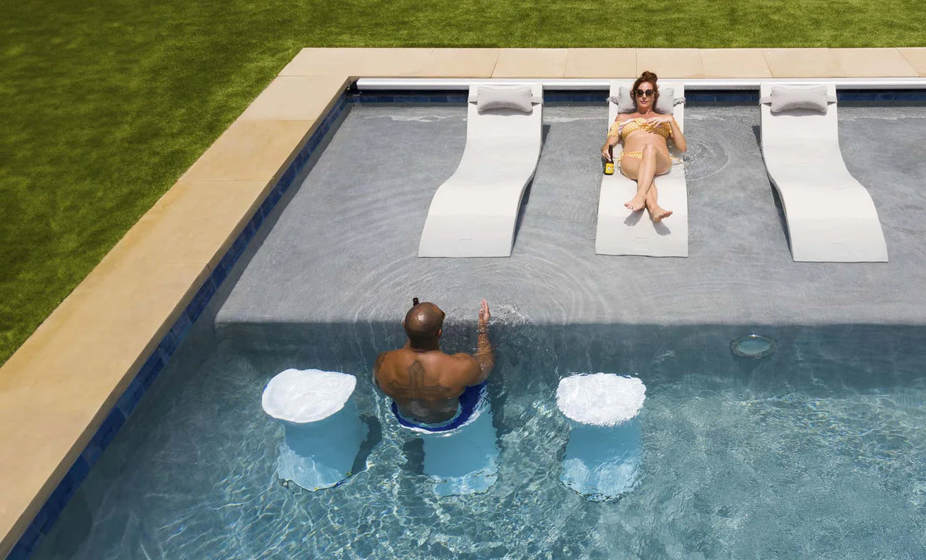 lady tanning on the shallow end of a pool while laying on ledge lounger chaise and man is sitting on pool edge on a bar height pool stool