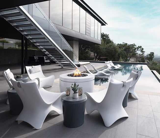 Contemporary outdoor patio with a round fire pit, surrounded by white modern chairs, a glass staircase, and a swimming pool overlooking a scenic view.