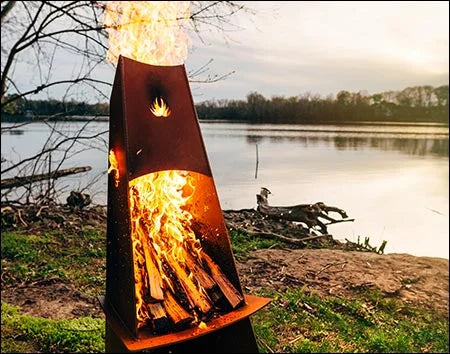 A tall, conical wood-burning fire pit with flames burning inside. The fire pit is surrounded by green grass and is near a body of water. There is a deer logo on the side of the fire pit.