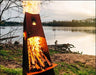 A tall, conical wood-burning fire pit with flames burning inside. The fire pit is surrounded by green grass and is near a body of water. There is a deer logo on the side of the fire pit.