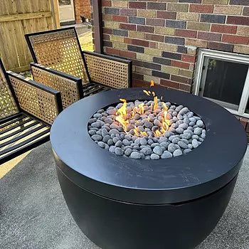 Round black fire pit lit with a flame, surrounded by gray stones and with two tan wicker chairs in the background on a paved patio.